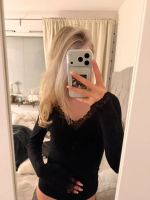 Svart långärmad topp med spets - Säljer en svart långärmad topp med v-ringning och fina spetsdetaljer vid halsringningen och ärmsluten. Toppen har en slim passform och är tillverkad i ett mjukt, stretchigt material som sitter snyggt på kroppen.