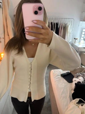 Krämvit kofta med knappar - Säljer en krämvit kofta med v-ringning och dekorativa tygklädda knappar framtill. Koftan har långärmad passform och är gjord i ett mjukt stickat material.