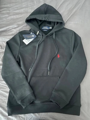 Svart hoodie från Polo Ralph Lauren - Svart hoodie från Polo Ralph Lauren med klassisk huva och dragsnören. Har en röd broderad logga på bröstet och en stor magficka framtill. Tillverkad i mjukt material som känns skönt mot huden. Perfekt för en avslappnad och stilren look.