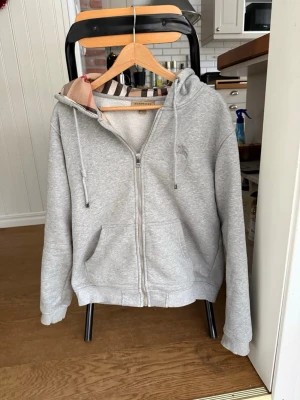 Grå zip hoodie från Burberry - Snygg grå hoodie från Burberry med dragkedja, huva med klassiskt rutigt foder och dragsko. Broderad Burberry-logga på bröstet. Tillverkad i mjuk bomullsblandning och har två fickor framtill. Perfekt för en chill och stilren look.
