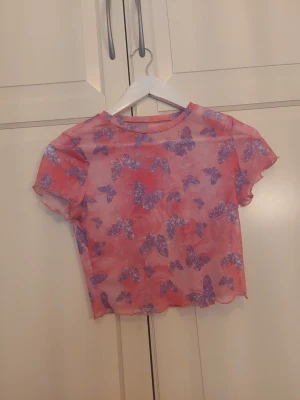 Rosa mesh topp med lila fjärilar - Säljer en transparent rosa mesh topp med lila fjärilsmönster. Toppen är croppad och har korta ärmar. Perfekt för dig som vill ha en söt och trendig look med lite extra färg och mönster.