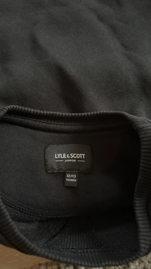 Svart sweatshirt från Lyle & Scott - En stilren svart sweatshirt från Lyle & Scott Junior. Tröjan har rund halsringning, ribbade muddar vid ärmslut och nederkant samt en klassisk passform. Perfekt för en avslappnad och trendig look.