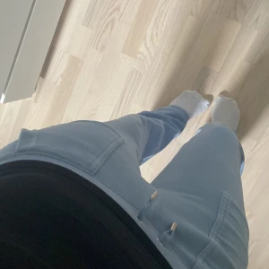 Ljusblå mjukisbyxor Juicy Couture - Supermjuka ljusblå mjukisbyxor från Juicy Couture med snörning i midjan och stora fickor med lock och tryckknappar. Klassisk avslappnad passform och riktigt skön kvalitet, perfekta för chill dagar.