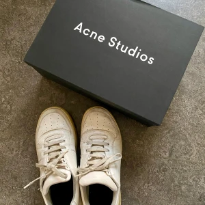 Acne Studios Steffey Lace Up Sneakers - Säljer ett par vita sneakers från Acne Studios med klassisk minimalistisk design. Skorna är i skinn med snörning, rund tå och den ikoniska smiley-detaljen bak på hälen. Perfekta för dig som gillar stilrena och trendiga skor. Begagnade men i fint skick. Originalkartong medföljer. 