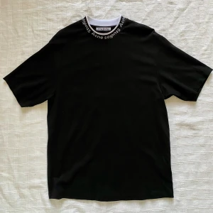 Acne Studios Logo Mock Neck Tee S NYSKICK - Svart t-shirt från Acne Studios med vit och svart ribbad krage med loggan invävd runt halsen. T-shirten är kortärmad och har en clean, stilren look. Materialet är en mix av viskos, polyamid och elastan för en mjuk och stretchig känsla. Använd endast en gång. Nypris: 2700kr
