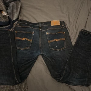 Mörkblå jeans från Nudie Jeans - Snygga mörkblå jeans från Nudie Jeans med klassisk orange söm och vågig brodyr på bakfickorna. Jeansen har fem fickor, knappgylf och är tillverkade i kraftigt denimtyg. Perfekta för dig som gillar stilrena och tidlösa jeans.W 30 L 30