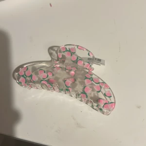 Hårklämma med rosa persikomönster - Superfin genomskinlig hårklämma dekorerad med söta rosa persikor och gröna blad. Perfekt accessoar för att piffa upp frisyren och ge en lekfull touch till din look. Klämman är i plast och har ett färgglatt, trendigt mönster.