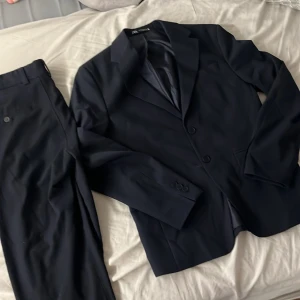 Mörkblå kostym från Zara, strl 44/ S-M - Snygg mörkblå kostym från Zara i storlek 44. Klassisk kavaj med två knappar, smala slag och matchande kostymbyxor med knapp och hällor för bälte. Kostymen har en stilren och modern passform, perfekt för dig som gillar en clean look.