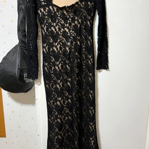 Svart lång spetsklänning med blommönster - Supersnygg långklänning i svart spets med blommigt mönster. Klänningen har lång ärm och figurnära passform, perfekt för dig som vill sticka ut. Materialet är genombrutet och ger en elegant look.