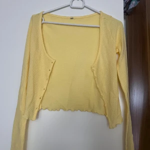 Gul croppad bolerotopp med knappar - Söt ljusgul bolerotopp med lång ärm och croppad passform. Toppen har små knappar framtill och vågig kant nertill för en extra detalj. Perfekt att slänga över ett linne eller klänning för en fräsch look.