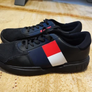 Tommy Hilfiger skor  - Tommy Hilfiger sneakers – nästan nya Snygga svarta sneakers från Tommy Hilfiger med klassiska röd, vit och marinblå detaljer på sidan. Skorna har snörning, mesh framtill och en bekväm platt sula. Nästan oanvända – bara provade några gånger, i riktigt fint skick.