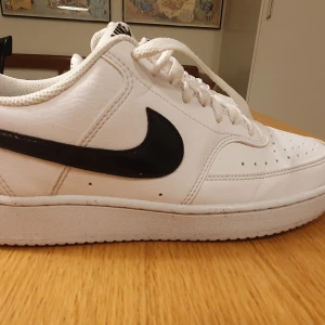 Nike Court Vision vit/svart EUR 40 - Klassiska Nike Court Vision sneakers i vitt skinn med svart swoosh-logga på sidorna. Lågt skaft, perforerad tå och vit platt sula. Skorna har snörning och är perfekta för dig som gillar en clean och tidlös look. Väldigt lite använda, så bra skick.