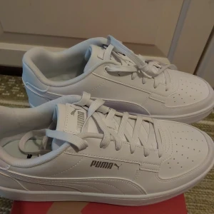 Vita sneakers från Puma - Fräscha vita sneakers från Puma med klassisk design. Skorna har snörning, perforerad tå och diskret Puma-logga på sidan. Ovandelen är i syntetmaterial och sulan är platt och greppvänlig. Perfekta för en clean och stilren look. Använd en gång.