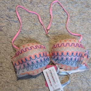 Färgglad balconette bikiniöverdel - Säljer en snygg balconette bikiniöverdel från Hunkemöller med vågigt mönster i rosa, blått och beige. Den har justerbara rosa axelband och guldfärgade detaljer. Perfekt för strandhäng och poolparty! (159 kr från början)☺️