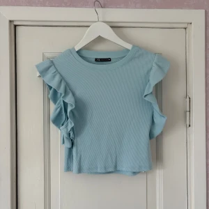 Ljusblå volangtopp från Zara - Supersöt ljusblå topp från Zara med volangdetaljer längs axlarna. Toppen är ribbad och ärmlös, vilket ger en fräsch och trendig look. Perfekt att styla med jeans eller kjol. 