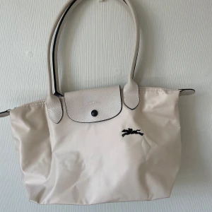 Beige/vit handväska från Longchamp - Snygg beige/vit handväska från Longchamp med klassisk design. Väskan har svarta detaljer, präglat lock i skinn och broderad logga framtill. Tillverkad i slitstark polyester med handtag i matchande färg. Perfekt storlek för vardagens essentials. Ny pris 1500, köpte fel storlek 
