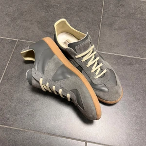 Maison Margiela gats - Maison Margiela sneakers i grå mocka och skinn med beige snörning och gummisula. Klassisk low-top siluett med rund tå och diskret branding på sulan. Tillverkade i Italien, stilrena och passar perfekt till streetwear eller casual outfits.