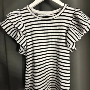 Randig volangtopp från Zara - Superfin topp från Zara med svarta och vita ränder. Modellen har korta volangärmar och rund halsringning. Ribbad struktur i tyget ger en snygg och trendig känsla. Inte använd många gånger!💓