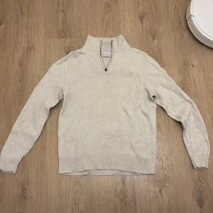Beige stickad half zip-tröja - Beige stickad tröja från These glory days med half zip-dragkedja i halsen. Tröjan har lång ärm, ribbade muddar och en avslappnad passform. Perfekt för lager-på-lager under kyliga dagar. Dock ett litet slitage på höger armbåge annars är den i mycket bra skick.