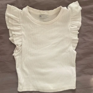 Vit volangtopp från Zara - Superfin vit topp från Zara med ribbad struktur och volangdetaljer på axlarna. Ärmlös modell som ger en söt och trendig look. Den är i mycket fint skick, använd ett fåtal gånger!