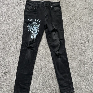 AMIRI jeans  - Svarta jeans från Amiri med skinny passform, slitna detaljer och tryckt ängelmotiv samt logga på vänster lår. Klassisk femficksmodell med silverfärgade metalldetaljer och läderpatch bak. Snygg streetstil med cool edge.