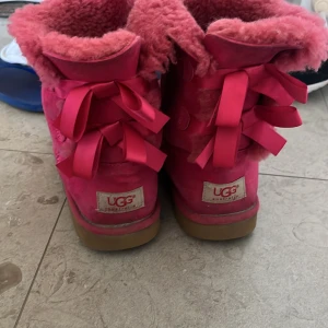 Rosa UGG boots med rosetter - Säljer ett par fluffiga rosa UGG boots med stora matchande rosetter baktill. Skorna har mjukt foder och ovandel i mocka, perfekt för kalla dagar. Snygg och mysig design som sticker ut med sin starka färg och detaljer.
