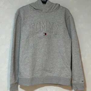 Grå hoodie från Tommy Hilfiger - Snygg grå hoodie från Tommy Hilfiger med broderad logga och klassisk flagga på bröstet. Tröjan har huva, långa ärmar och ribbade muddar. Perfekt för en chill och stilren look. 
