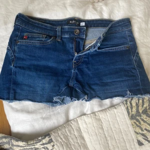 Blå jeansshorts från Big Star, strl 27 - Snygga blå jeansshorts från Big Star med push up-effekt. Klassisk femficksmodell, rå kant nedtill och detaljerade sömmar. Shortsens midja är normal och de har dragkedja och knapp framtill. Perfekt för dig som gillar en casual och trendig look.