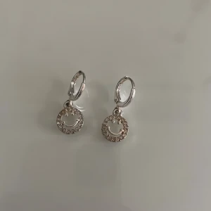 Silverörhängen med smiley och stenar - Ett par snygga örhängen i silverfärg med hängande smiley-ansikten dekorerade med små glittriga stenar runt kanten. Perfekt för dig som vill ha en trendig detalj till din look.