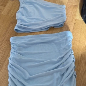 Ljusblått sätt med rynkade detaljer - Snyggt ljusblått sätt med one shoulder-topp och kort kjol. Båda delarna har rynkade detaljer som ger en trendig look. Perfekt för en dag ute i värmen. Enkel och stilren design som sticker ut.