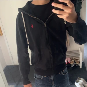 Svart hoodie från Polo Ralph Lauren XS - Svart hoodie från Polo Ralph Lauren i storlek XS. Klassisk design med vit snörning i huvan, dragkedja framtill och röd broderad logga på bröstet. Mjuk bomullskänsla och känguruficka framtill. Perfekt för en chill och stilren look.