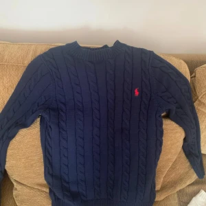 Marinblå stickad tröja från Ralph Lauren - Snygg marinblå stickad tröja från Ralph Lauren med klassiskt kabelstickat mönster och röd logga på bröstet. Rund hals och långa ärmar, perfekt för dig som gillar stilren och tidlös design.