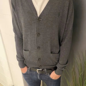 Grå kofta från Polo Ralph Lauren - Snygg grå kofta från Polo Ralph Lauren med klassisk v-ringning och knappar framtill. Mjuk bomullskvalitet, två fickor och diskret logga broderad på bröstet. Perfekt att slänga över en t-shirt för en chill och stilren look.                         Passar M