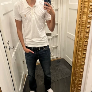 Vit pikétröja från Polo Ralph Lauren - Klassisk vit pikétröja från Polo Ralph Lauren med broderad logga på bröstet. Tröjan har korta ärmar, krage och knappar framtill. Tillverkad i mjuk bomull som känns skön mot huden. Perfekt för en clean och stilren look.