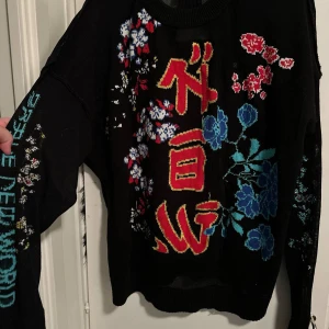 Svart stickad tröja med asiatiska tecken strl: - Unik svart stickad tröja med färgglada asiatiska tecken och blommigt mönster i rött, blått, vitt och gult. Tröjan har lång ärm, öppen rygg och coola broderade detaljer på både fram- och baksida. Perfekt för dig som vill sticka ut med din stil.