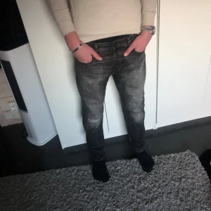 Jack n Jones jeans - 29/32 men sitter som 32/32. Säljer ett par svarta jeans med snygga slitningar och tvättad look. Jeansen har klassisk femficksdesign, raka ben och normal passform. Materialet är robust denim och färgen är mörkgrå/svart med slitna detaljer på framsidan för en cool vibe.
