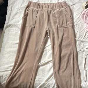 Beiga mjukisbyxor Juicy Couture L - Supermjuka beiga mjukisbyxor från Juicy Couture i storlek L. Byxorna har bred resår i midjan, stora fickor fram och bak med snygga silverknappar och diskret logga. Perfekta för chill dagar och har en klassisk Juicy-look i glansig velour.