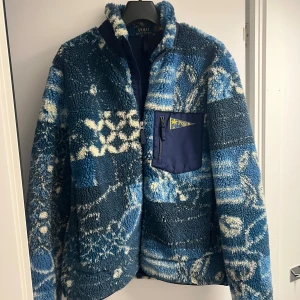 Ralph Lauren fleecejacka - Säljer en blå fleece från Polo Ralph Lauren med coolt mönster i vitt och mörkblått. Jackan har en bröstficka med dragkedja och Polo-logga, samt hög krage. Materialet är mjuk polyester och akryl, perfekt för kalla dagar.