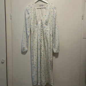 Vit långklänning med blå blommor från Adoore - Supersöt långklänning från Adoore i vit satin med ljusblå blommigt mönster. Klänningen har v-ringning, långa ärmar och knappar framtill. Endast använd ett fåtal gånger,därför u mycket bra skick!🩵