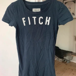 Mörkblå t-shirt Abercrombie & Fitch XS - Snygg mörkblå t-shirt från Abercrombie & Fitch i storlek XS. Klassisk rund hals och korta ärmar. Stor vit text 'FITCH' framtill ger en cool college-vibe. Mjuk bomull som är skön mot huden och passar perfekt till jeans eller shorts.