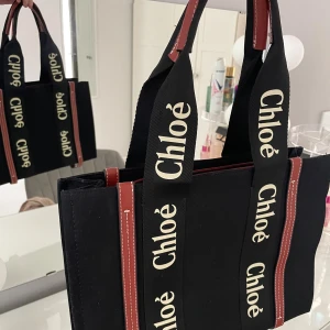 Svart axelväska Chloé med logoband - Snygg svart axelväska från Chloé med tydliga logoband i beige och svarta färger. Väskan har bruna detaljer med vita sömmar och är tillverkad i bomull och polyester. Rymlig insida med dragkedjefack och praktiska handtag. Perfekt för dig som gillar statement-accessoarer.