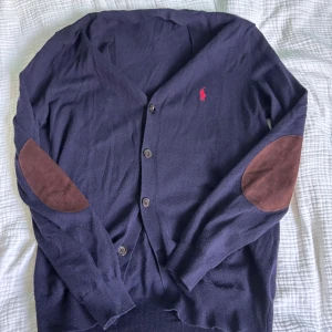 100% merino ull mörkblå kofta från Polo Ralph Lauren - Mörkblå kofta från Ralph Lauren med klassisk röd logga på bröstet. Tröjan har knappar framtill, långa ärmar och bruna mockadetaljer på armbågarna. Tillverkad i 100% merino ull material.