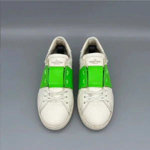 Valentino Garavani Open sneakers vit/grön - Snygga Valentino Garavani Open sneakers i vitt skinn med en grön bred rem över mitten. Klassisk låg modell med rund tå, vita snören och ikoniska nitar baktill. Perfekta för dig som vill sticka ut med en färgklick i din outfit.