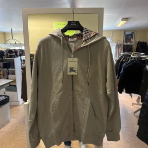 Beige hoodie från Burberry med dragkedja - Snygg beige hoodie från Burberry med klassiskt rutigt foder i huvan och dragkedja framtill. Tröjan har snörning i huvan, två fickor och broderad Burberry-logga på bröstet. Tillverkad i 100% bomull för en skön och avslappnad känsla.