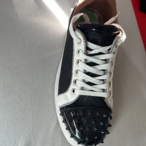 Loobsträffas bara upp i Skåne  - Unika sneakers i svart och vitt med glittrande detaljer och vita snören. Tåpartiet är täckt av svarta nitar för en edgy look. Skon har en klassisk röd sula och inslag av skinn och textil. Perfekt för dig som vill sticka ut med din stil.