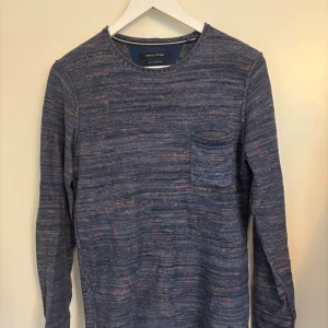 Blå stickad tröja Marc O'Polo - Säljer en blåmelerad stickad tröja från Marc O'Polo med diskreta inslag av orange och rosa. Tröjan har rund hals, bröstficka och lång ärm. Perfekt för dig som gillar en chill och stilren look. Liten logotyp nertill på ena sidan.