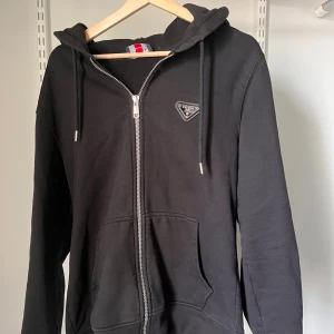 Prada hoodie - Svart hoodie från Prada med klassisk huva, dragkedja framtill och snörning. Tröjan har en snygg logotyp på bröstet och stora fickor framtill. Perfekt för dig som vill ha en stilren och lyxig streetwear-look.
