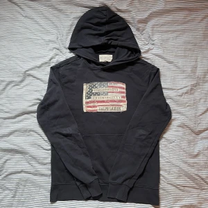 Denim & Supply Hoodie - Svart hoodie från Ralph Lauren Denim & Supply med stor amerikansk flagga i tyg på bröstet och broderade detaljer. Klassisk huva och lång ärm, perfekt för en avslappnad streetstil. Mjuk bomullskänsla och snygg vintage-look. STORLEK M