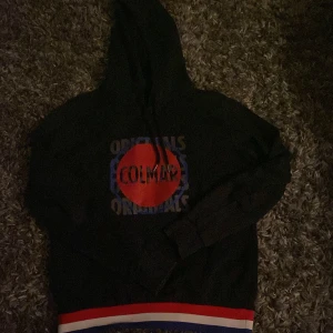 Svart Colmar hoodie med röd logga - Svart hoodie från Colmar Originals med stor röd logga på bröstet och text i blått och vitt. Tröjan har huva med snörning och ribbad nederkant med röd, vit och blå rand. Tillverkad i mjuk bomull, perfekt för chill dagar. Snygg och sportig vibe. Äkta de stor storlek 14y men skulle säga den sitter som xs