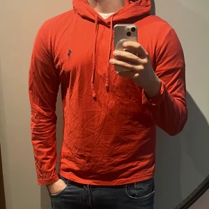 Ralph Lauren hoodie - Röd hoodie från Ralph Lauren med klassisk blå logga broderad på bröstet. Tröjan har huva med snörning och är tillverkad i mjuk bomull. Långärmad och med en avslappnad passform, perfekt för en chill look. Endast en minimal defekter som syns på sista bilden :)